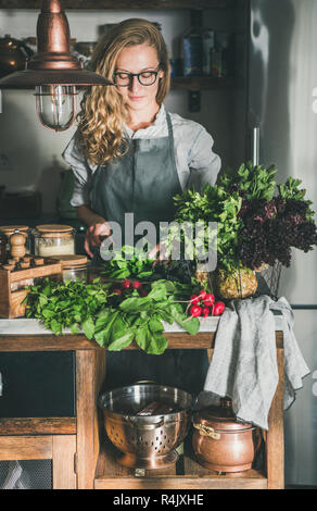 Herbst saisonalen vegetarischen, veganen Abendessen kochen. Junge Frau in Leinen Schürze und Gläser schneiden von Kräutern und Gemüse auf Beton Arbeitsplatte in der Küche. Langsam Stockfoto