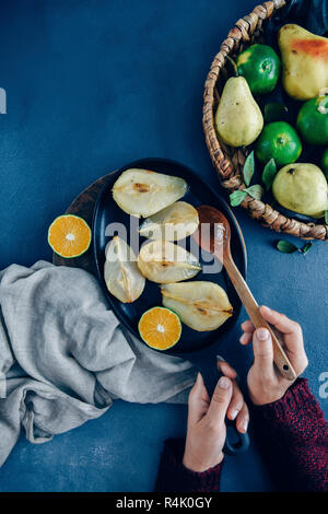 Hände halten einen Löffel aus Holz und eine eiserne Pfanne mit karamellisierten Birnen und einen Keil o orange Innen. Frische Birnen und Orangen in einem Korb auf der Seite. Stockfoto