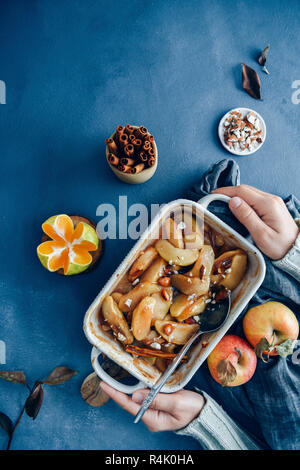 Hände halten Zimt gebacken Apfelscheiben in eine Backform von Äpfel, Orangen, Mandeln und Zimtstangen begleitet. Stockfoto