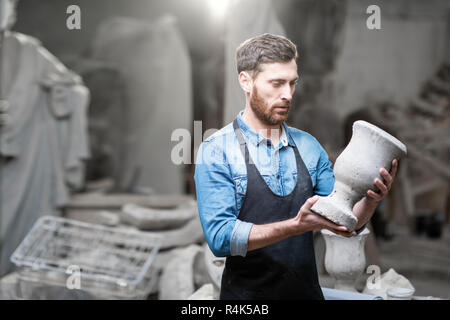 Porträt einer stattlichen Bildhauer in blauen T-Shirt und Schürze Holding alte Vase im Studio mit Skulpturen auf dem Hintergrund Stockfoto