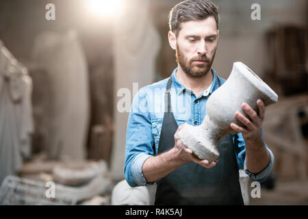 Porträt einer stattlichen Bildhauer in blauen T-Shirt und Schürze Holding alte Vase im Studio mit Skulpturen auf dem Hintergrund Stockfoto