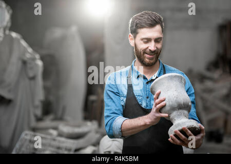 Porträt einer stattlichen Bildhauer in blauen T-Shirt und Schürze Holding alte Vase im Studio mit Skulpturen auf dem Hintergrund Stockfoto