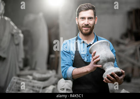 Porträt einer stattlichen Bildhauer in blauen T-Shirt und Schürze Holding alte Vase im Studio mit Skulpturen auf dem Hintergrund Stockfoto
