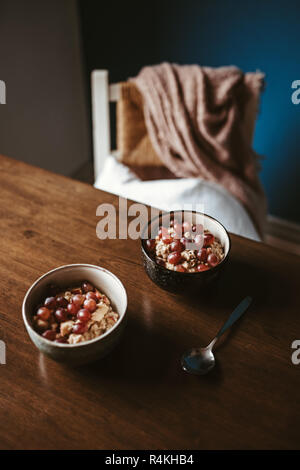 Zwei Schalen mit Porridge mit Trauben auf einen hölzernen Tisch mit Stuhl und Decke im Hintergrund Stockfoto