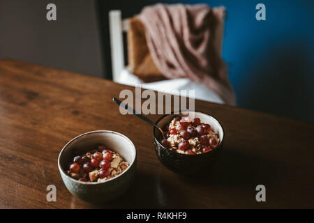 Zwei Schalen mit Porridge mit Trauben auf einen hölzernen Tisch mit Stuhl und Decke im Hintergrund Stockfoto