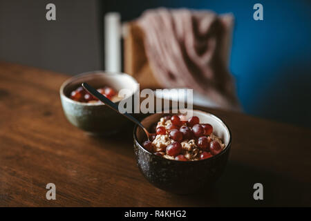 Zwei Schalen mit Porridge mit Trauben auf einen hölzernen Tisch mit Stuhl und Decke im Hintergrund Stockfoto