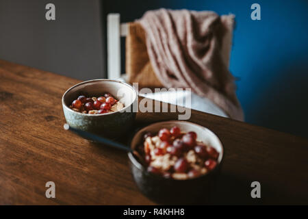 Zwei Schalen mit Porridge mit Trauben auf einen hölzernen Tisch mit Stuhl und Decke im Hintergrund Stockfoto