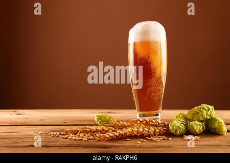 Schließen Sie ein Glas Bier mit Schaum und Blasen, grün Hopfen und Gerste Korn und Spikes auf hölzernen Tisch über dunkelbraunen Hintergrund mit Kopie Raum, Stockfoto