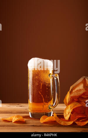 Schließen Sie ein Glas mug Lagerbier mit weißen Schaum und Blasen und Papier Tüte Kartoffelchips auf hölzernen Tisch über dunkelbraunen Hintergrund mit Kopieren s Stockfoto