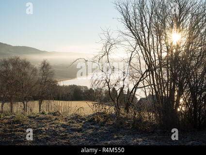 Landwirtschaftliche ländlichen Natur Landschaft mit Misty stimmungsvollen Blick über idyllische Ackerland, den See und die Berge bei Sonnenaufgang, mit Sonnenstrahlen durch die Bäume Stockfoto