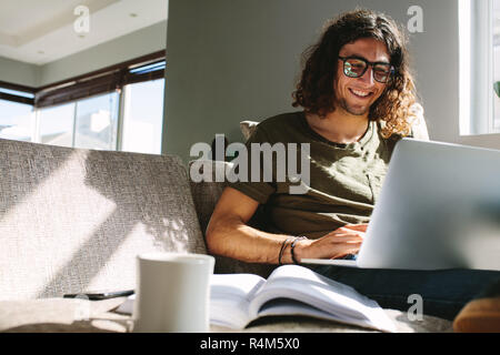 Lächelnd männlicher Student zu Hause sitzen und über einen Laptop. Schüler sitzen auf einer Couch studieren auf Laptop mit Büchern und Kaffee Tasse auf den Tisch. Stockfoto