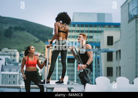 Fitness-Mann steht mit weiblichen Athleten, die eine Wasserflasche halten und auf dem Dach reden. Männer und Frauen in Fitnesskleidung, die danach eine Pause machen Stockfoto