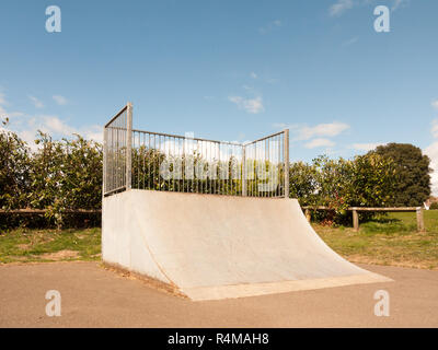 Eine leere und ungenutzte Ramp Halfpipe am Skatepark im Country Park in Großbritannien glänzen im Sonnenlicht des Tages und mit einem Strauch Hecke hinter den Bars es Schutz eingezäunt Stockfoto