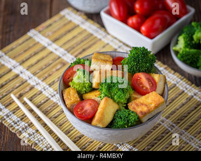 Gegrillter Tofu mit Brokkoli und Tomaten in Weiß Schüssel auf Bambus Pad mit Stäbchen auf der linken Seite. Stockfoto