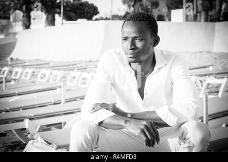 Fashion schwarz-weiß Bilder Portrait afrikanischen schönes Modell Mann sitzt am Strand auf der Suche vor ihm - Schönheit Konzept für attracti Stockfoto