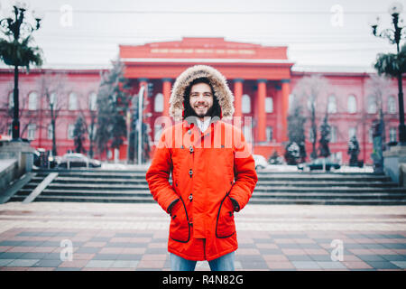 Porträt, Nahaufnahme der Jungen stilvoll Mann lächelnd mit einem Bart rot Winter Jacke mit Kapuze und Fell auf dem Kopf bekleidet steht gegen die backgro Stockfoto