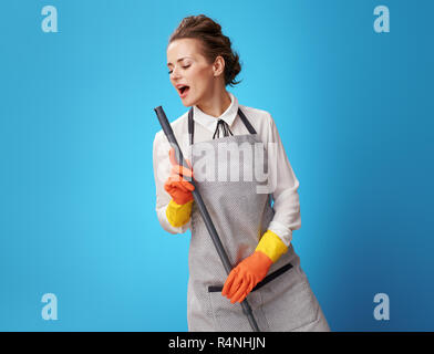 Glückliche junge Putzfrau in Schürze mit Mop singen auf blauen Hintergrund. Spaß und schnell ordentlich das Haus oder Büro Hausmädchen Reinigung. Stockfoto