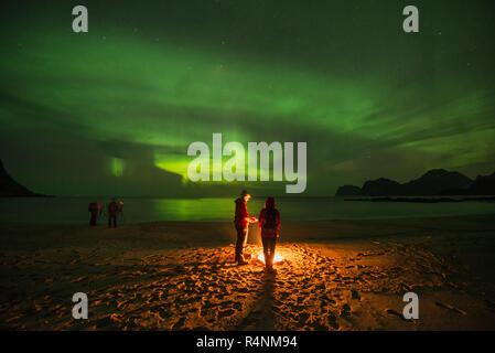 Ruhige Szene mit Menschen um Lagerfeuer am Strand unter auroraÂ borealisÂ bei Nacht, Flakstadoya, Lofoten, Norwegen Stockfoto