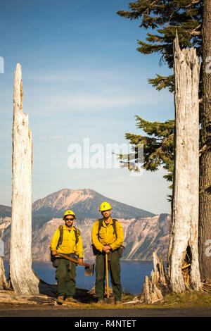 Zwei wildland Feuerwehrmänner in den gelben Hemden stehen mit ihrer Ausrüstung zwischen zwei tote Bäume mit Bergsee im Hintergrund. Crater Lake, Oregon, USA Stockfoto