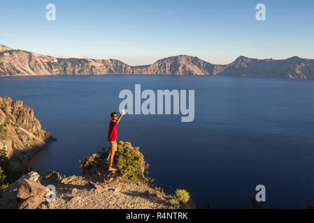 Ein junger Mann in einem roten T-Shirt stellt für eine selfie auf einem Felsvorsprung hoch über einem tiefblauen See umgeben von Bergen, Crater Lake, Oregon, USA Stockfoto