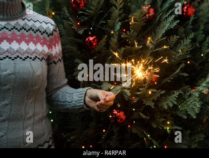 Frau in Form gestrickter Pullover Wunderkerzen halten vor dem Weihnachtsbaum Stockfoto