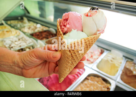 Weibliche Hand eine große Erdbeere, Minze, Vanille mit Sauerkirschen Eis in der Waffel Kegel. Eis Kühlschrank mit Stahl Service Container. Stockfoto