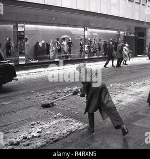 1960, historische, ein Mann im Mantel und trug eine Kappe mit einem Pinsel räumen Schnee von der Straße gegenüber der Kleidung Einzelhändler C&A, in der Nähe der Oxford Street, London, England, UK. Die C&A-Gebäude war Ihre erste Britische Shop und Handel in Großbritannien bis 2001. Stockfoto