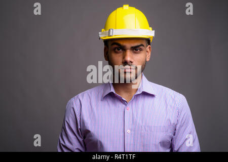 Junge hübscher indischer Geschäftsmann tragen, hardhat gegen Grau b Stockfoto