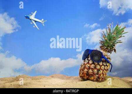 Ananas Obst mit Sonnenbrille auf dem Sandstrand zu einem fliegenden Flugzeug in den blauen Himmel mit Wolken suchen, Sommerferien Konzept, kopieren Raum Stockfoto