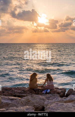 Zwei Frauen sitzen auf den Felsen bei Sonnenuntergang mit Blick auf das Meer, Tel Aviv, Israel Stockfoto