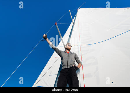 Junger Mann steht auf Yachtcharter Ausleger und freut sich. Er hält am Mast Seil mit Hand. Junger Mann stellt. Er trägt graue Hoody und schwarze Hosen. Stockfoto