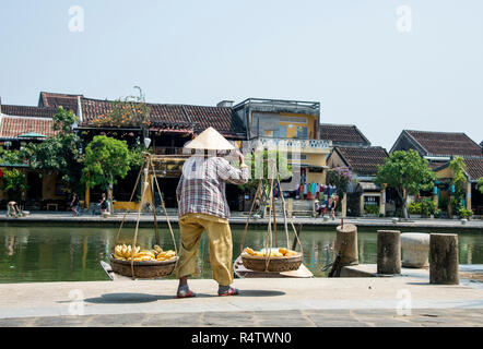 Hoi An einem typischen alten vietnamesischen Dame eine Schulterpasse, beladen mit Obst und Gemüse entlang einer Straße in der Altstadt von Hoi An Stadt Vietnams. Stockfoto