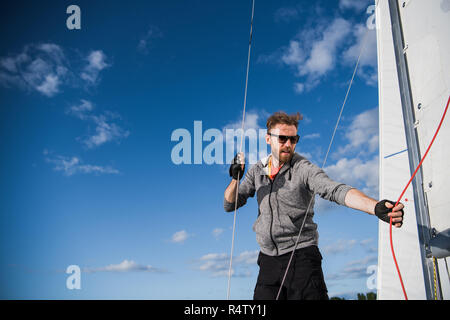 Gut aussehender bärtiger Matrose arbeiten mit Seilen an Deck einer Yacht gegen den klaren, blauen Himmel und Meer Stockfoto