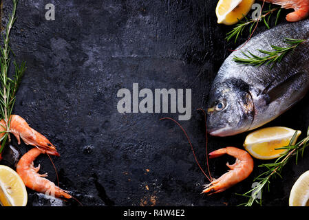 Blick von oben auf die frische Meeresfrüchte auf grunge texturiertem schwarzen Hintergrund. Garnelen, Brassen, Kräuter, Zitrone und Eis auf Schiefer mit Copyspace. Stockfoto