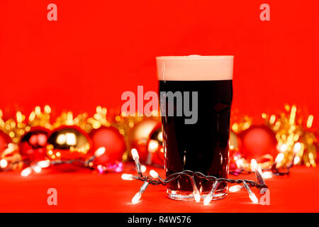 Nonik pint Glas dunkles Bier oder stout Ale mit Weihnachtsbeleuchtung Christbaumkugeln und Lametta auf rotem Hintergrund Stockfoto