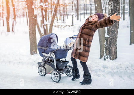 Junge schöne Mutter in einem pelzmantel Tanzen beim Spaziergang mit einem Kinderwagen in einem Stadtpark im Winter. Glückliche Mutterschaft Konzept. Genießen Sie unbeschwerte Mater Stockfoto