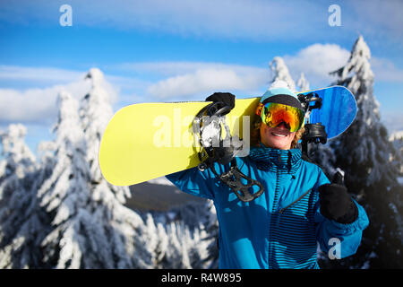 Lächelnd posiert Snowboard Snowboarder Tragen auf den Schultern am Ski Resort in der Nähe von Forest vor Freeride Session. Reiter Daumen oben Zeichen tragen polarisierte Brille. Moderne Snowboardausrüstung. Stockfoto