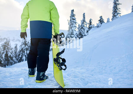 Rückansicht der Snowboarder Klettern mit seinem Brett auf dem Berg für backcountry Freeride Session im Wald. Mann mit Snowboard Wandern am Ski Resort. Reiter lime modische Outfit. Stockfoto