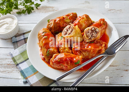 In der Nähe von kohlrouladen gefüllt mit Hackfleisch und Reis auf einem weißen Teller auf einem alten rustikalen Tisch mit saurer Sahne in einer Schüssel serviert, Ansicht von oben Stockfoto