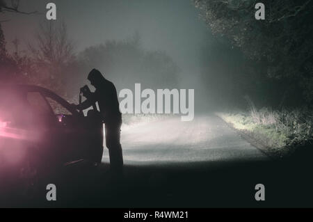 Ein einsamer hooded Abbildung in ein Auto suchen. Auf eine gespenstische Silhouetted, Land nebligen Winter Straße bei Nacht. Mit einem kalten, Körnigen bearbeiten. Stockfoto