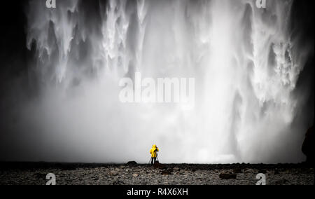 Ein einsamer Fotograf in Gelb steht an der Basis der Skogafoss Wasserfall an der südlichen Küste von Island Stockfoto
