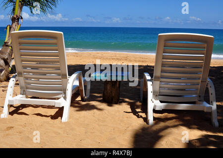 Zwei weißen Liegestühlen am Meer mit einem kleinen blauen Tisch dazwischen. Stockfoto