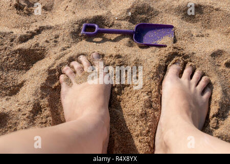 Eine enge ebf Blick auf Frauen Füße mit einem kleinen violetten Spaten vor für sie auf dem Strand Sand an einem sonnigen Sommertag Stockfoto