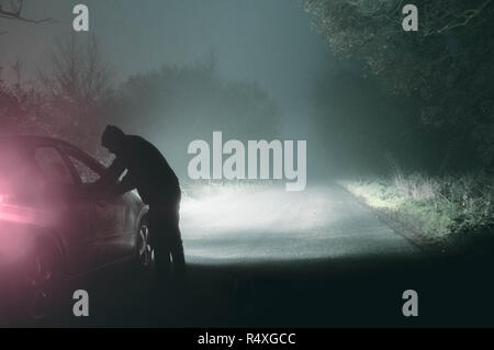 Ein einsamer hooded Abbildung in ein Auto suchen. Auf eine gespenstische Silhouetted, Land nebligen Winter Straße bei Nacht. Mit einem kalten, Körnigen bearbeiten. Stockfoto