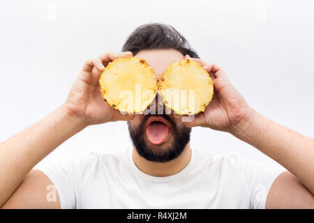 Lustig und positiven jungen Mann stehen und decken Sie die Augen mit zwei Hälften einer Ananas. Auch er die Zunge auf Kamera zeigen. Auf weissem Hintergrund. Stockfoto