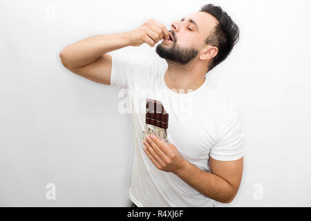 Bild voller Freude. Junge bärtige Mann steht und isst Schokolade. Er setzt Stück davon in den Mund. Kerl hält die Augen geschlossen. Man ist konzentriert. Auf weissem Hintergrund. Stockfoto