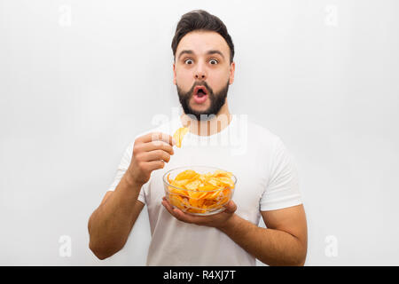 Aufgeregt und junger bärtiger Mann stehen und Blick in die Kamera begeistert. Er hält Schüssel mit Chips in der einen Hand und einem Stück von ungesunden Produkt in ein anderes. Auf weissem Hintergrund. Stockfoto