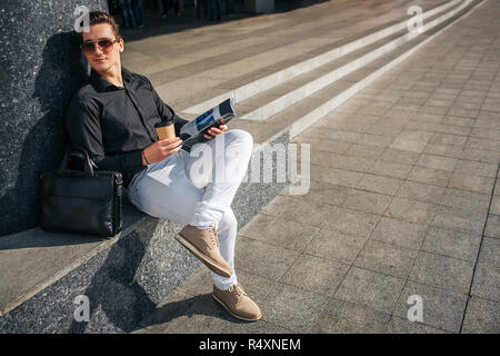 Junger Mann auf die Schritte sitzen und durch die Brille schauen Sie nach rechts. Er hält Tasse Kaffee und Journal. Kerl hat ein Bein nach dem anderen. Stockfoto