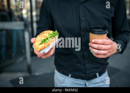 Schnittansicht der Mann hält salzig croissan und Tasse trinken. Er steht an Gebäude. Kerl trägt schwarze T-Shirt und eine blaue Jeans. Stockfoto