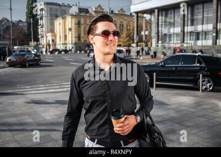 Schöne und glückliche junge Mann auf der Straße. Er schaut nach rechts durch eine Sonnenbrille. Junge Mann hat Tasse trinken und Ledertasche. Er ist zuversichtlich. Stockfoto